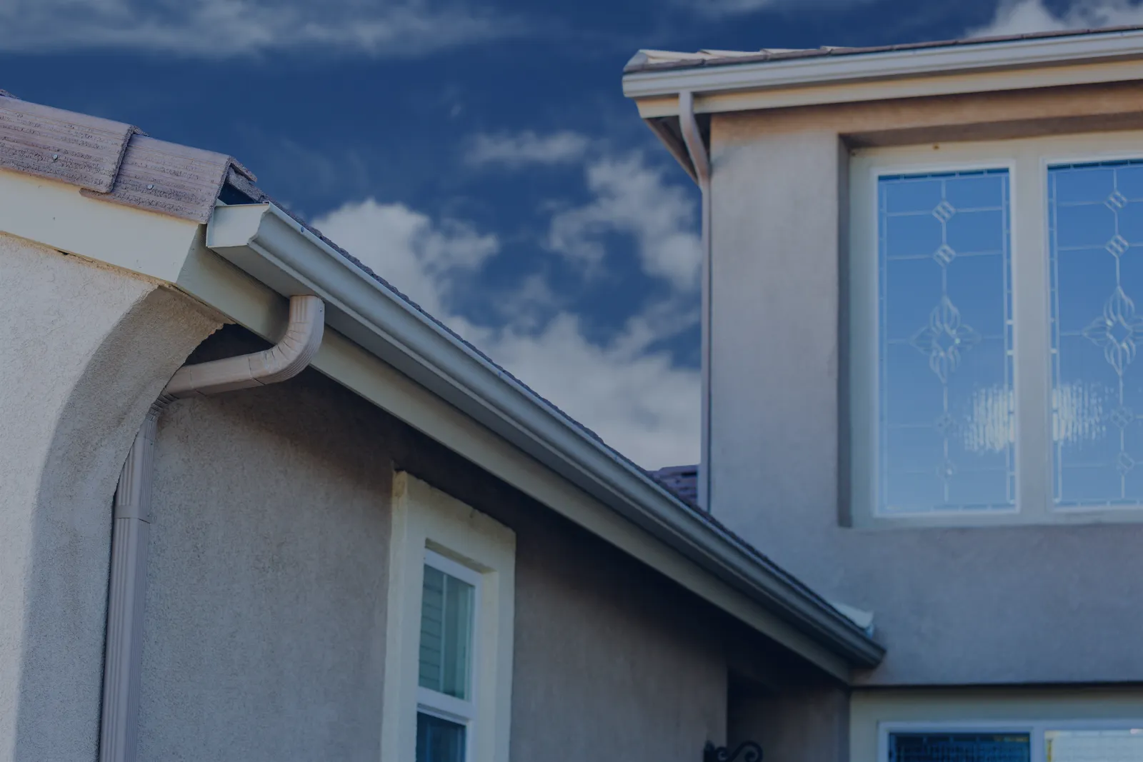 Close-up of a metal rain gutter running along the eave of a tan stucco two-story house.