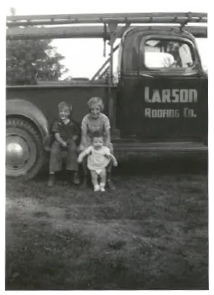 Three children standing in front of a vintage truck with the sign 'Larson Roofing Co.' on its door.