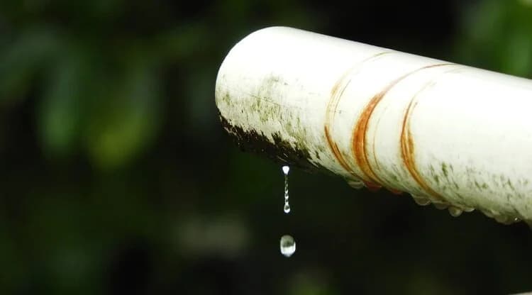 A close-up of a weathered white gutter downspout pipe with a single water droplet dripping from its end against a dark green background.