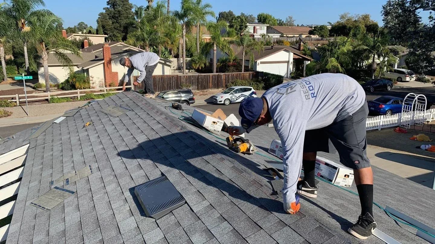 Two roofers fastening gray asphalt shingles on a residential roof.