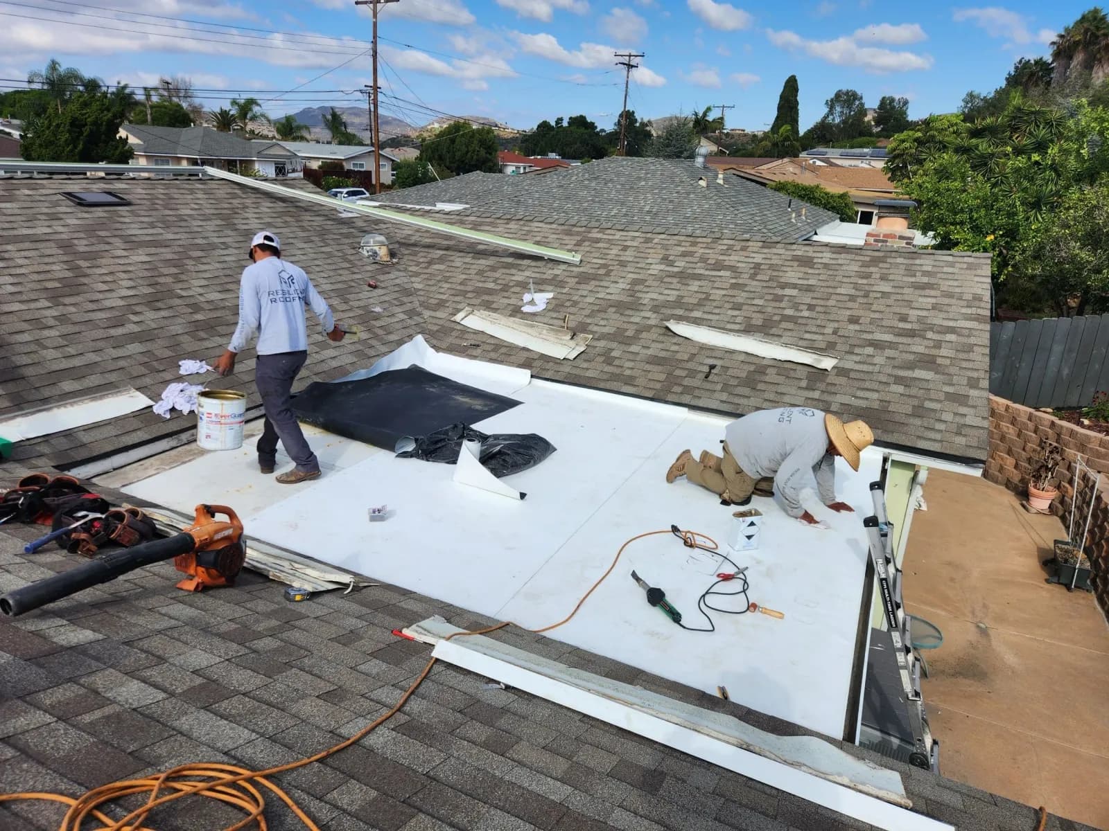 Two roofing workers installing a white flat-roof membrane on a residential roof.