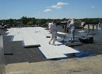 Workers installing white membrane sheeting on a large flat commercial rooftop.