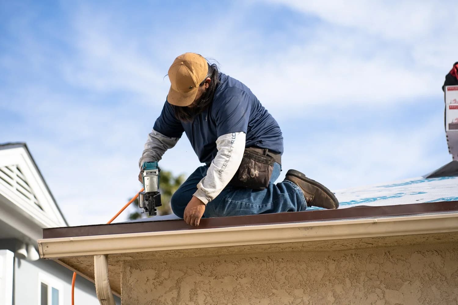 A roofer kneeling at a roof eave using a pneumatic nail gun while installing shingles over synthetic underlayment.