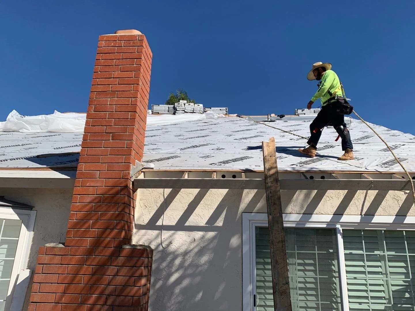 Roofer in safety gear working on a roof covered with white underlayment next to a brick chimney.