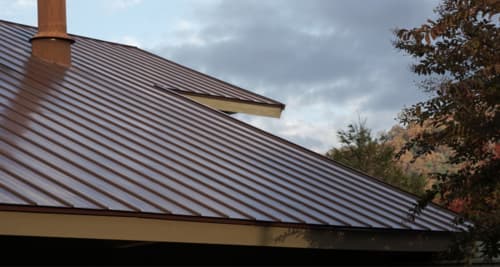A brown copper standing-seam metal roof with a chimney pipe against a cloudy sky.