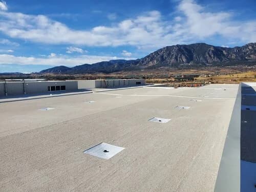 A large white flat commercial membrane roof with drains, set against a desert landscape and mountains under a blue sky.