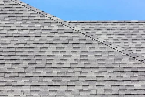 Gray architectural shingles meeting at a roof ridge line against a clear blue sky.