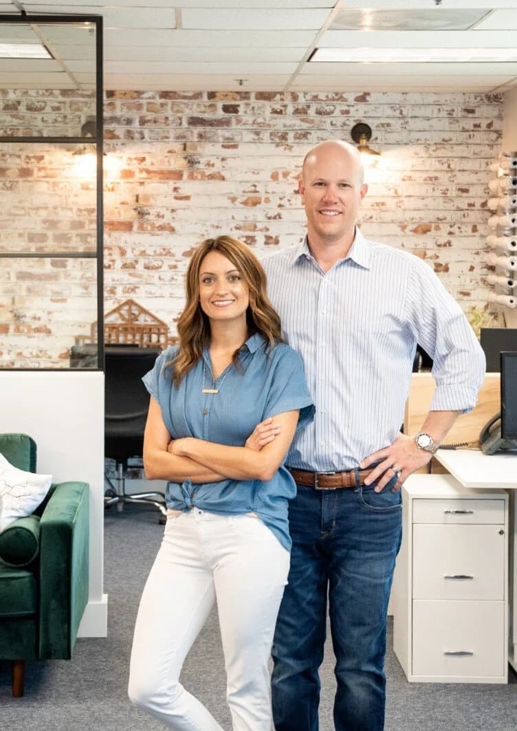 A woman and a man standing together in an office, smiling at the camera. The woman has shoulder-length brown hair, wearing a blue short-sleeve blouse and white pants. The man is taller, bald, wearing a light blue striped button-up shirt and dark jeans.