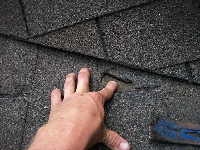 A hand points to a cracked and torn asphalt shingle on a roof next to a pry bar.