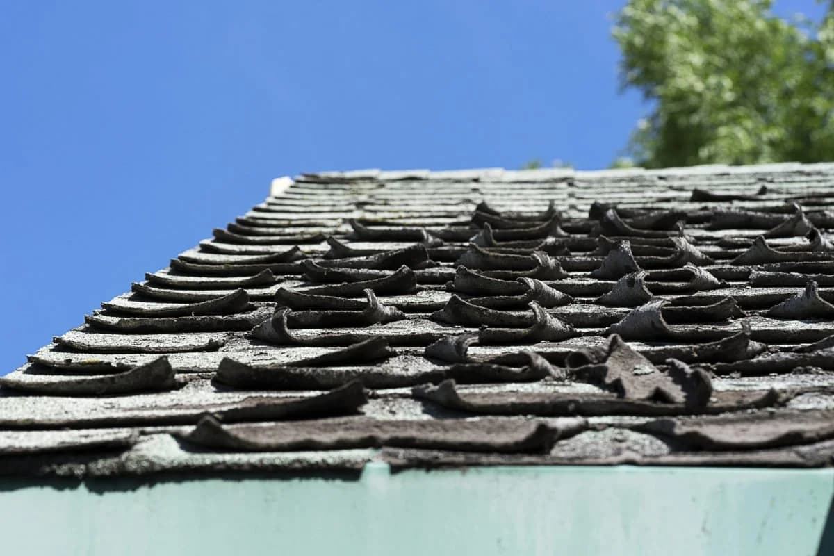 Severely curled, warped roof tiles against a blue sky, showing heavy weather damage.