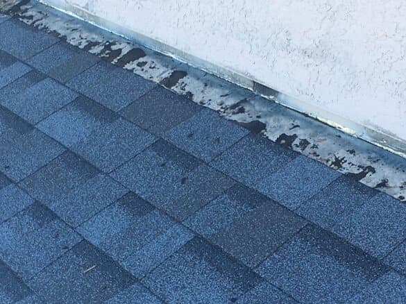 A close-up of gray asphalt shingles meeting a stucco wall with deteriorated, peeling flashing along the joint.