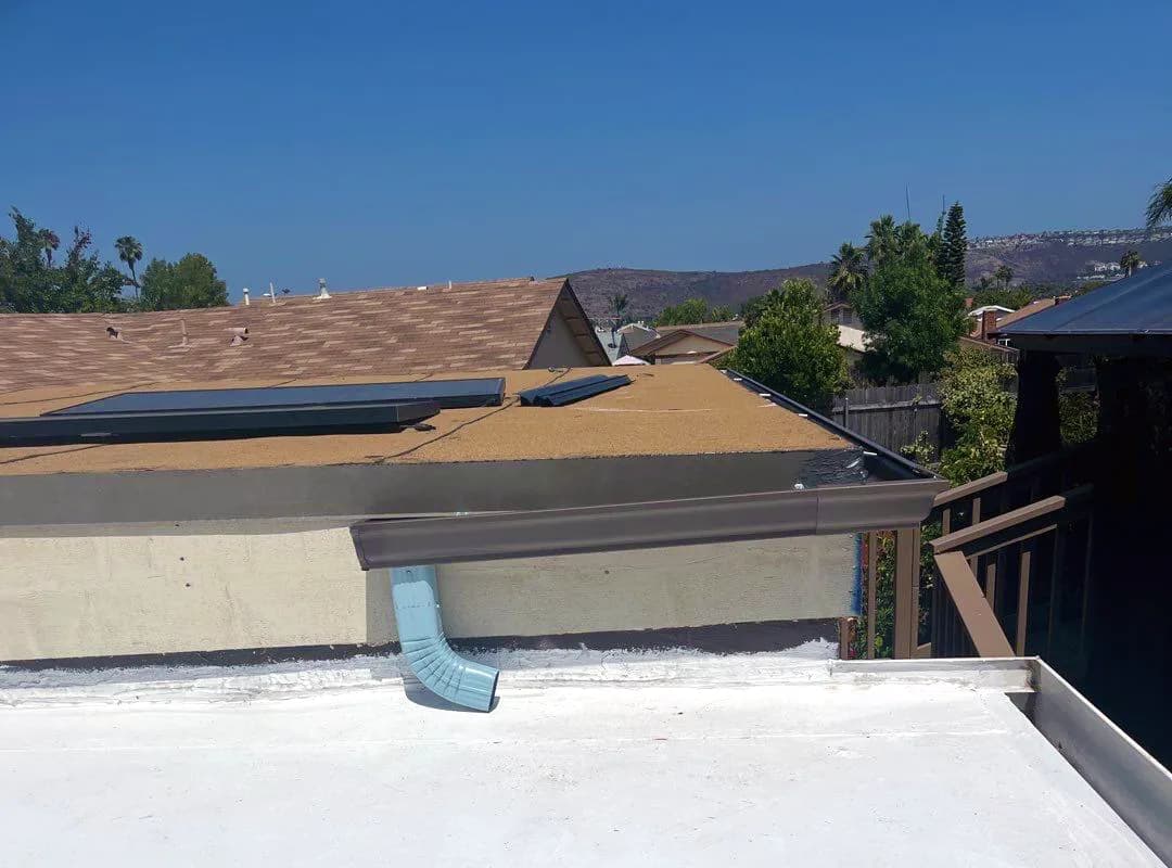 A white-coated flat roof section with a metal downspout and adjacent roofs behind.