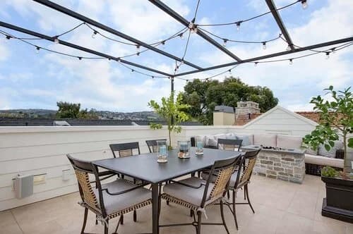 A rooftop terrace with a dining table, string lights, and built-in seating overlooking a hilly neighborhood.