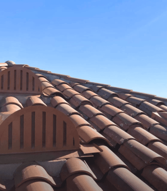 Two tile-matched louvered Brandguard dormer vents set into a Spanish clay tile roof against a clear blue sky.