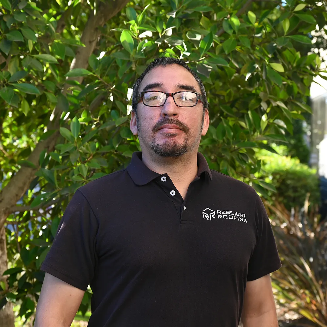 Man with glasses and facial hair standing outdoors in front of green leafy bushes, wearing a black polo shirt with 'Resilient Roofing' logo.