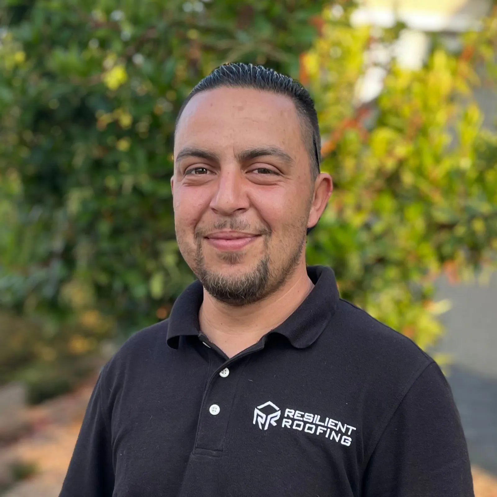 A man with dark hair and a goatee, smiling, wearing a black polo shirt with the logo and text "Resilient Roofing," standing outdoors with green and yellow foliage in the background.