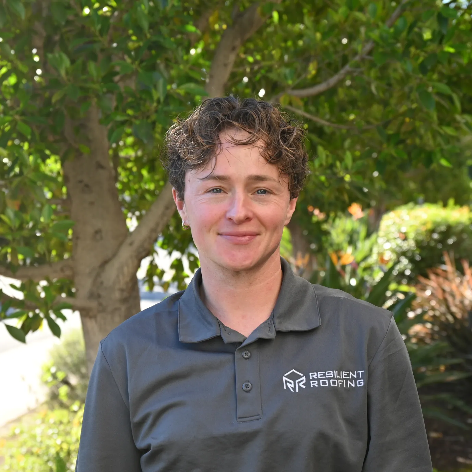 Close-up of a person wearing a dark gray shirt with a logo and text for 'Resident Roofing' on the chest, standing outdoors with a large green leafy tree in the background, smiling gently.