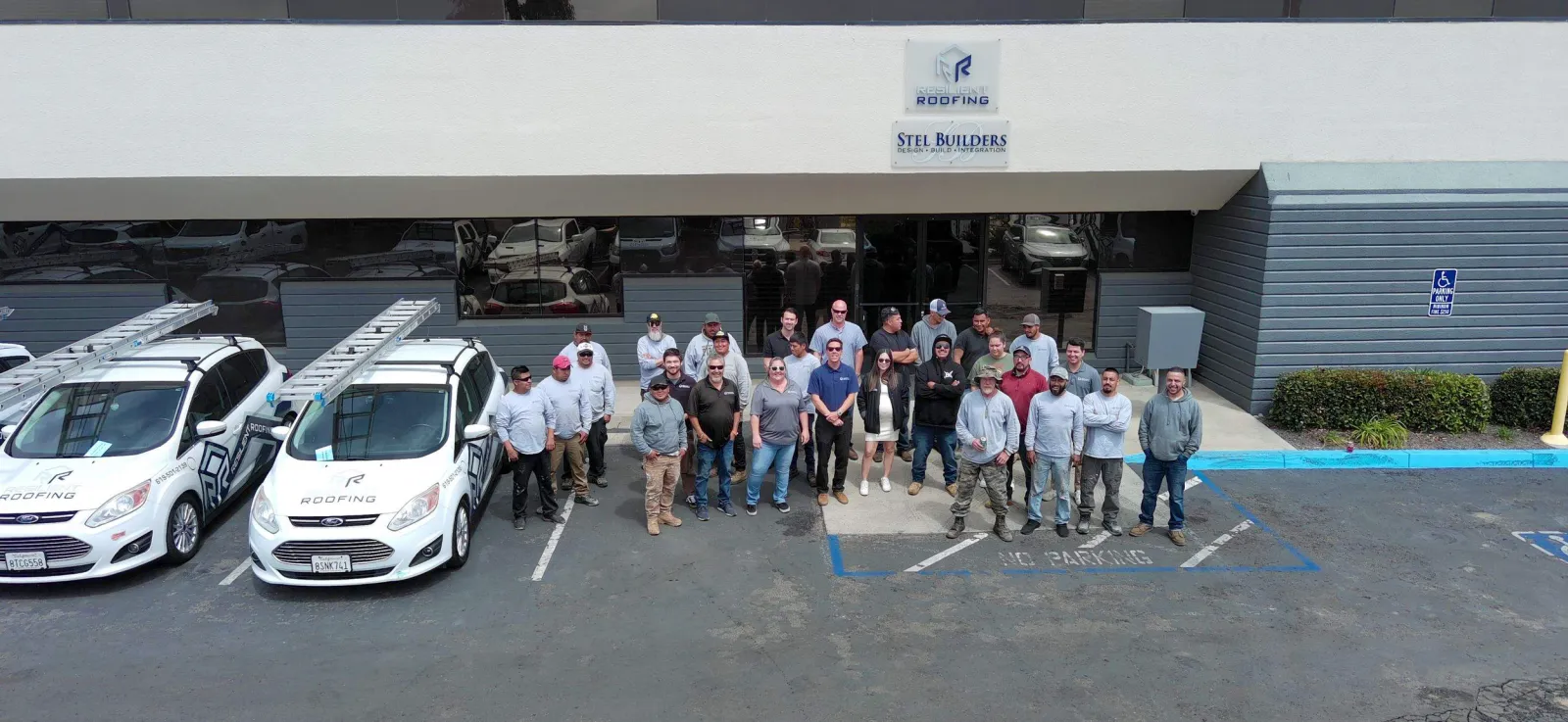 A group of people standing in front of a commercial building named 'Stel Builders' and 'Resilient Roofing' with parked cars including two white company vehicles with ladders on top, and a blue parking space labeled 'No Parking'.