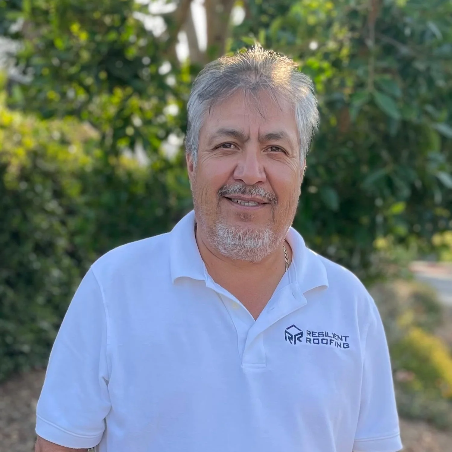 A middle-aged man with gray hair and a beard smiling outdoors. He is wearing a white polo shirt with a logo that says 'Resilient Roofing' on it. Green trees and sunlight are in the background.