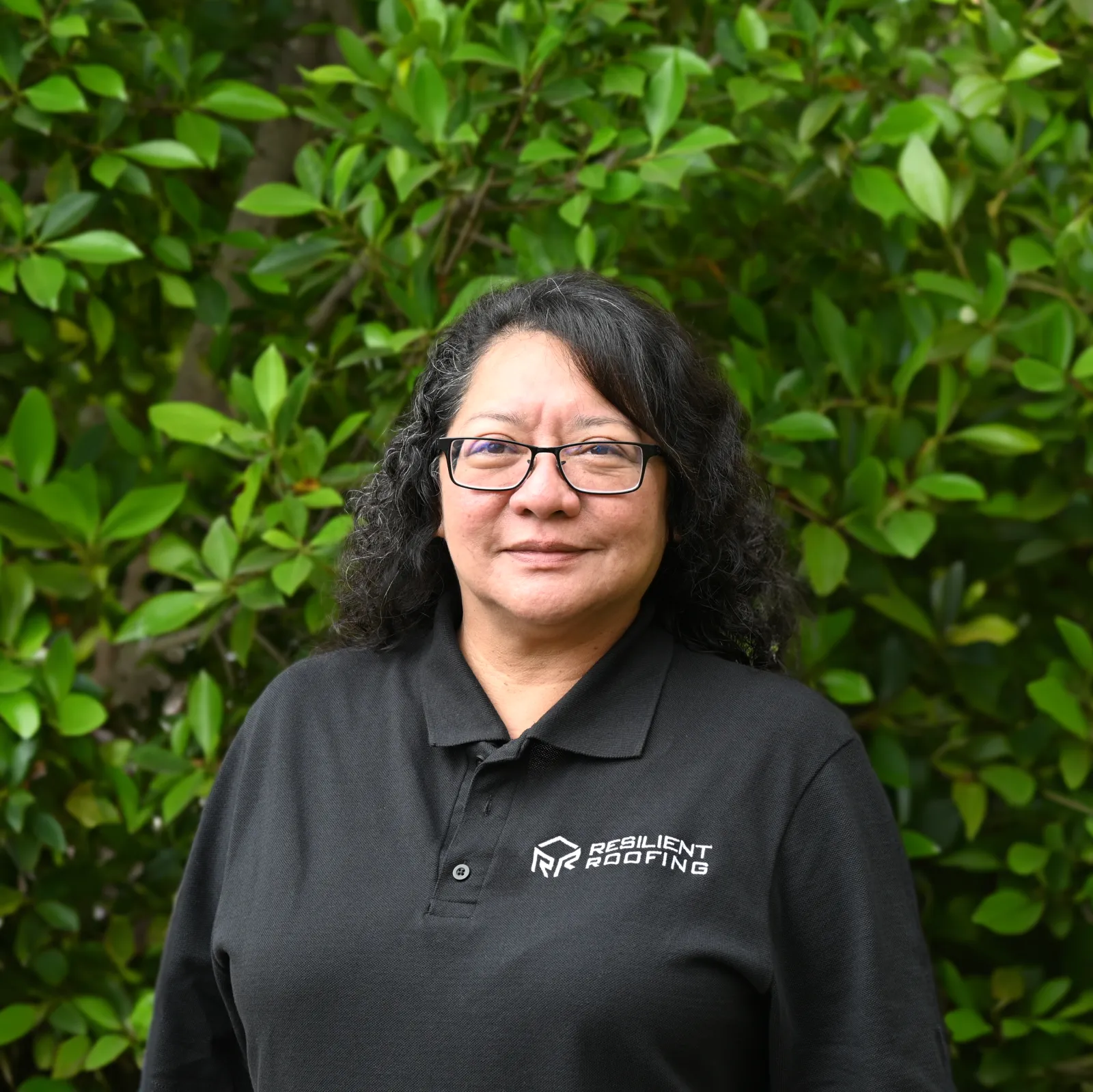Close-up of a woman with curly black hair and glasses, wearing a black polo shirt with a logo that reads 'Resilient Roofing', standing in front of green bushes.