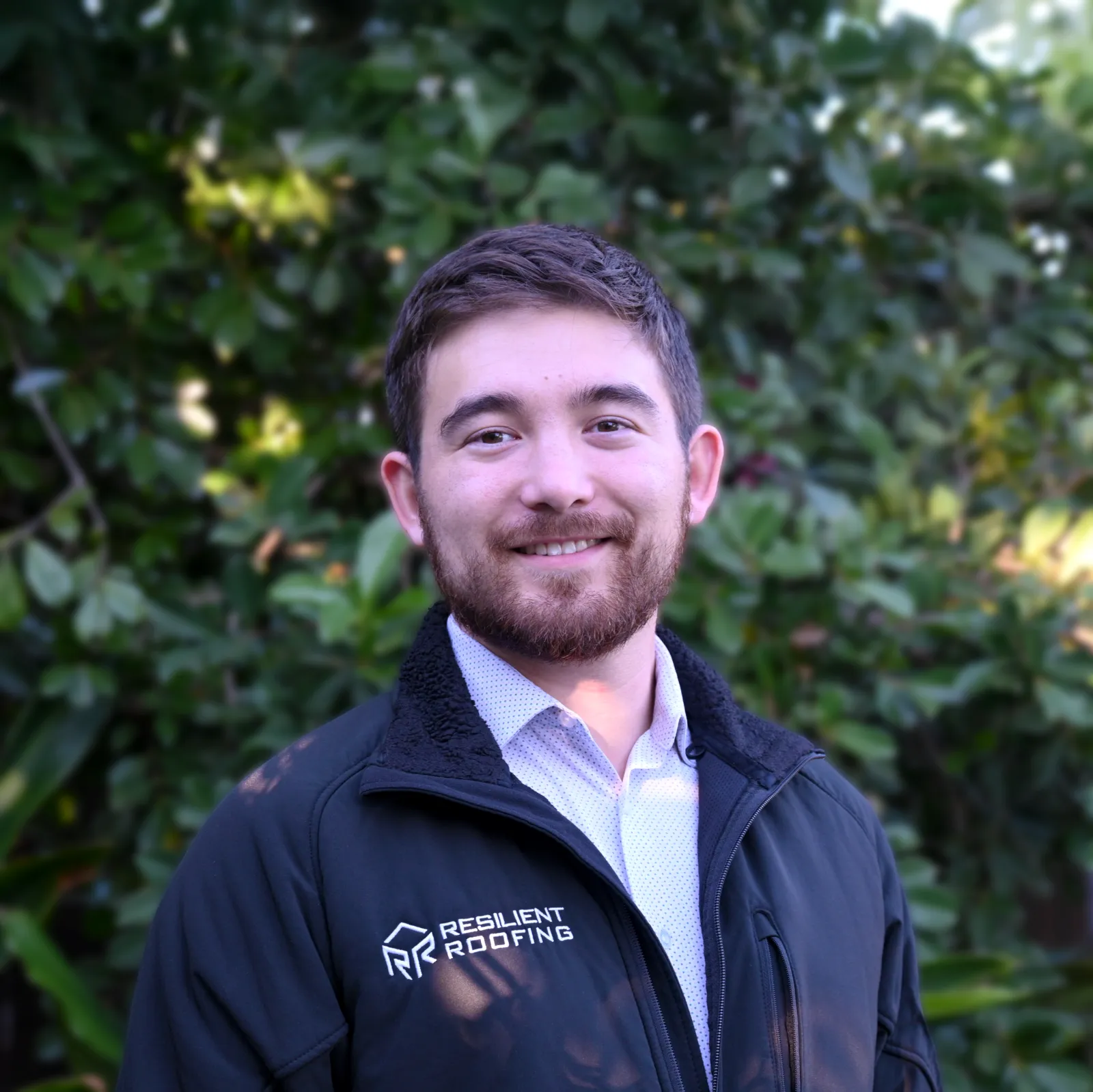 A young man with short brown hair and a beard smiling, wearing a black jacket with the logo and text 'Resilient Roofing' on it, standing outdoors in front of green leafy bushes.