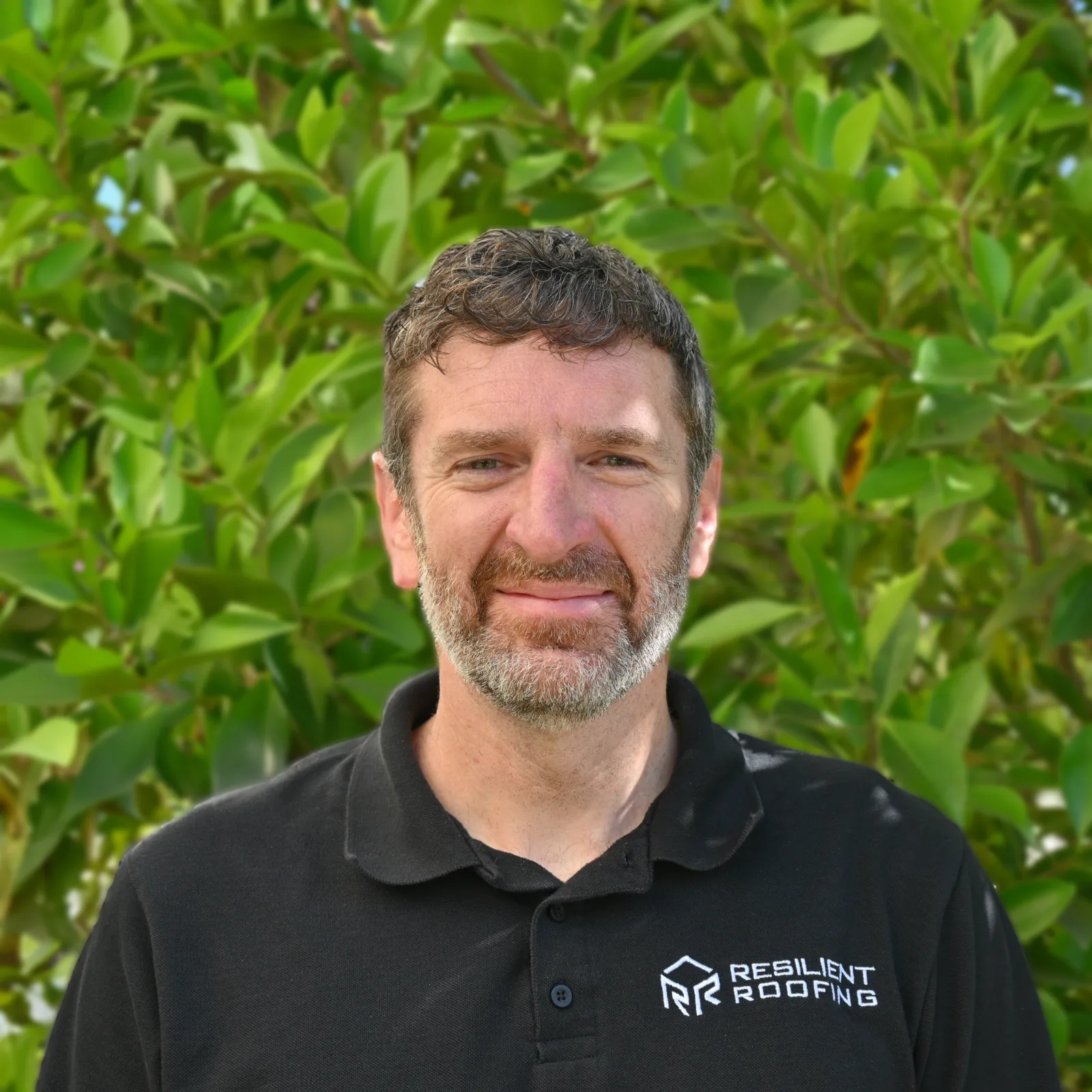 Close-up of smiling man with dark curly hair, wearing a black polo shirt with a corporate logo, standing in front of green foliage.