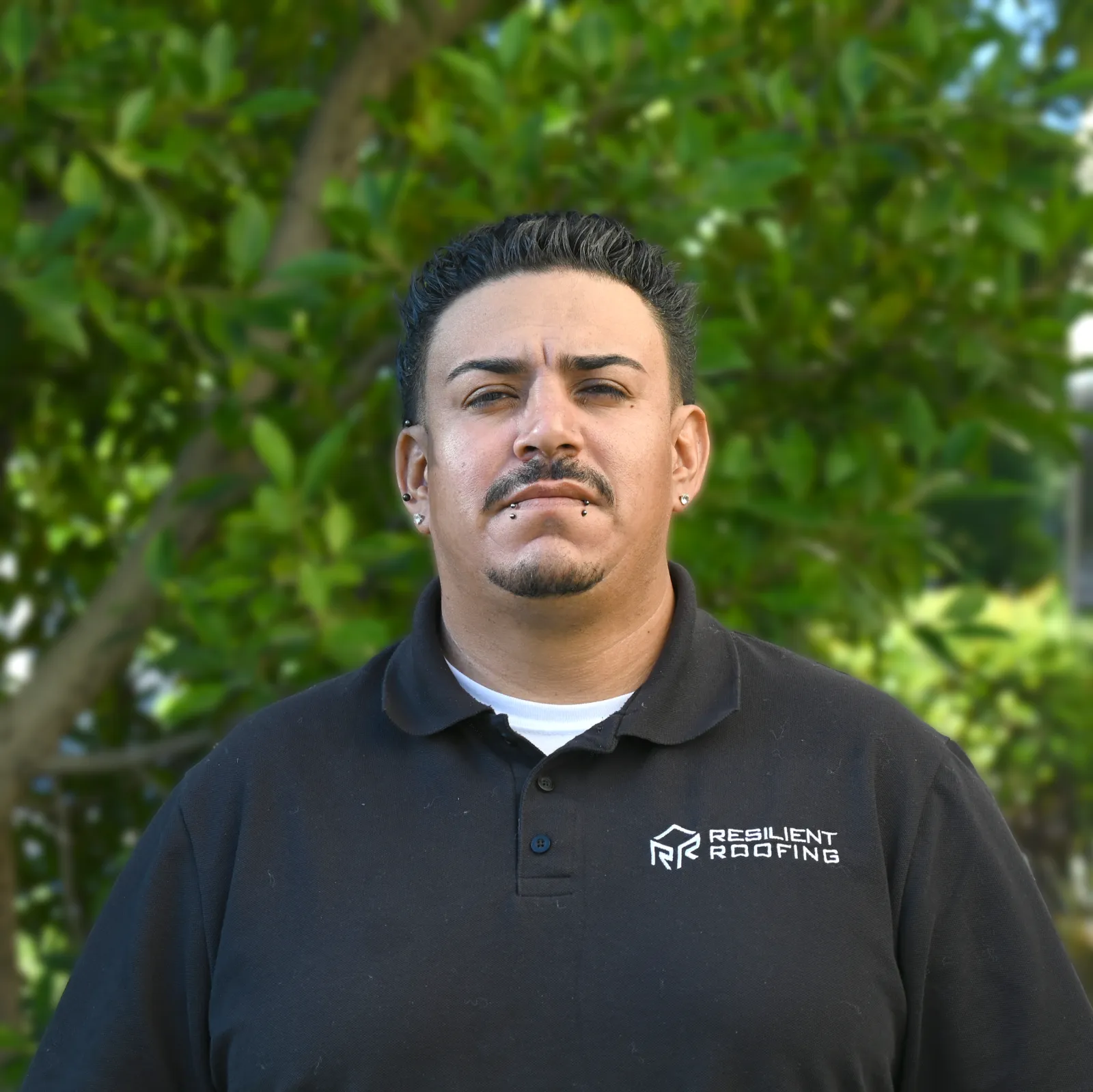 Close-up of a man wearing a black polo shirt with 'Resilient Roofing' logo, standing outdoors with green trees in the background.