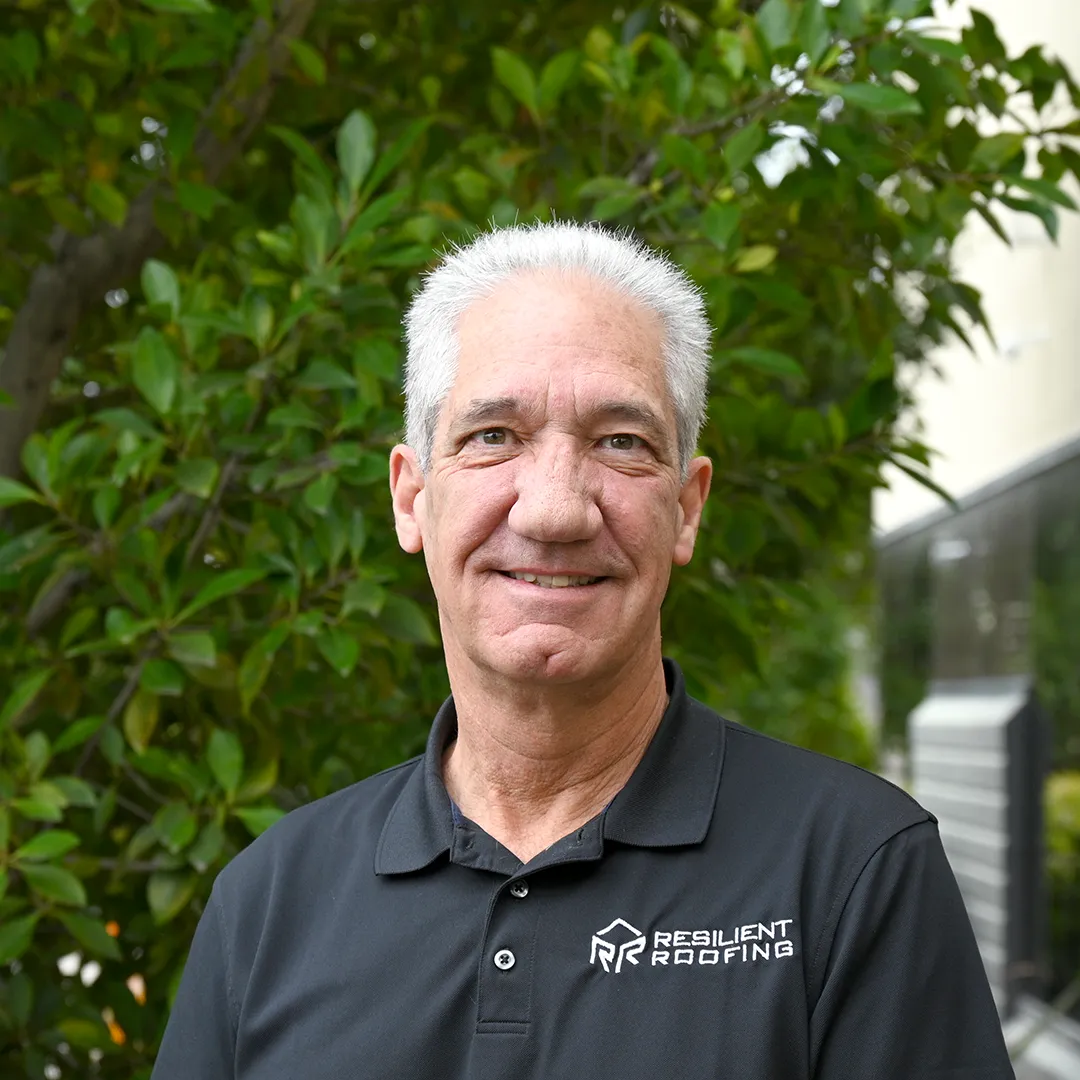 A smiling older man with gray hair wearing a black polo shirt with 'Resilient Roofing' logo, standing outdoors in front of green bushes.