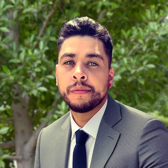 A man with dark hair and a beard wearing a gray suit, white shirt, and black tie, standing outdoors in front of green tree foliage.